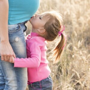 girl in pink sweater and grey jeans kissing tummy of pregnant woman in blue shirt and blue denim jeans