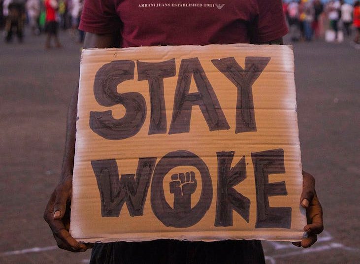 man wearing a hat holding a placard with a text stay woke